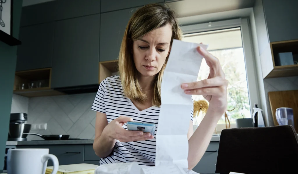 A lady looking upset as she looks at a bill and tries to calculate finances on her phone. 