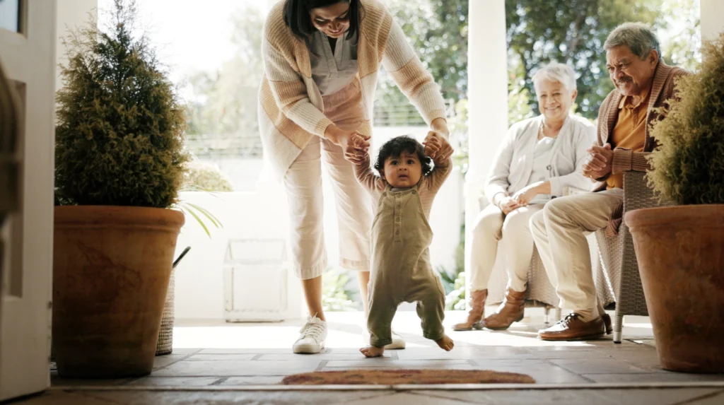 A baby walking with his mother while doting grandparents look on. 