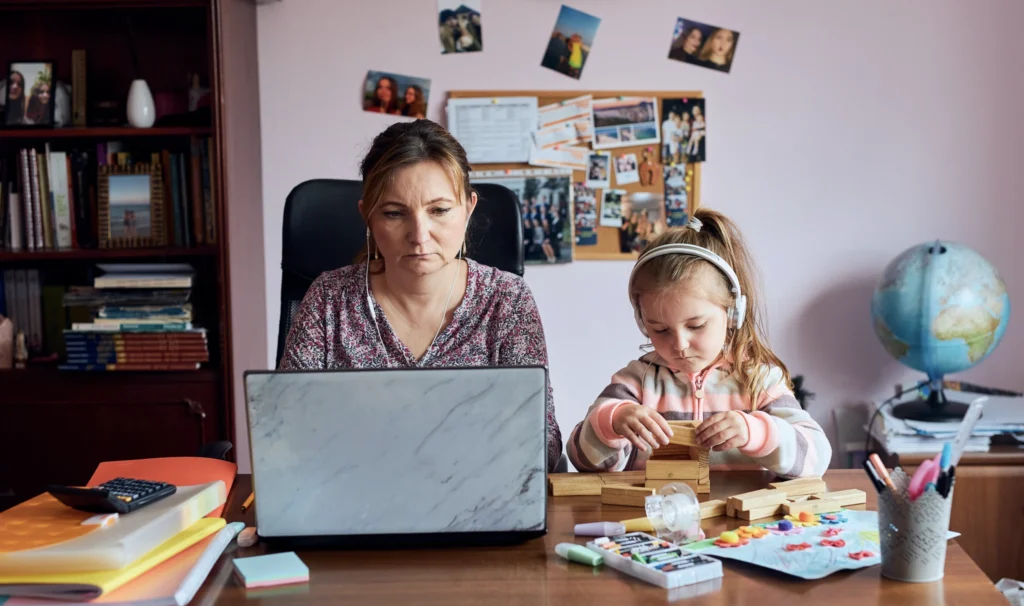 A mother working on her computer with her daughter beside her, playing